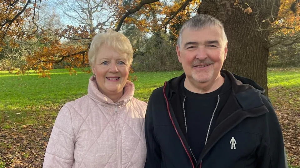 Tony Collier, who is white, and his wife Tracey are standing on a field in a park. It is an autumn day and the leaves have fallen from the trees. 