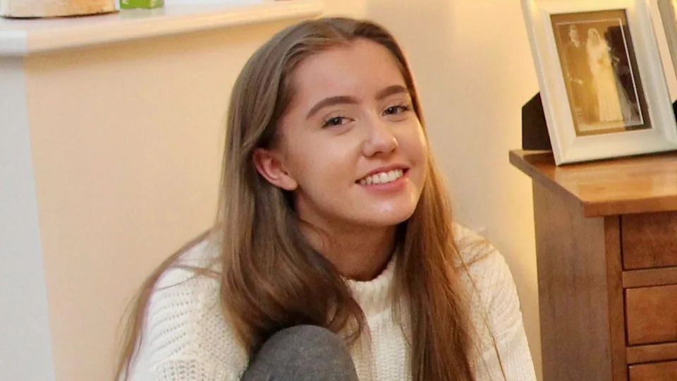 Emily smiles for the camera. The picture is taken inside a house and she is sat on the floor, with her back resting against the wall. She has long, light-brown hair.