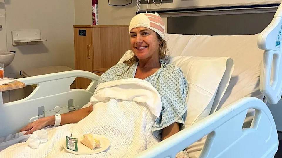 A woman in a hospital bed with her head bandaged, she is wearing a hospital gown and smiling. 
