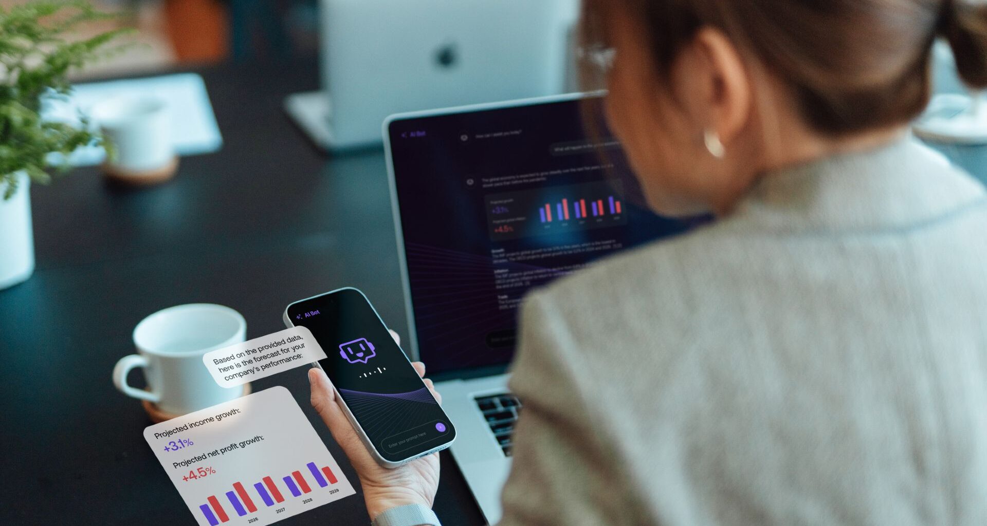 A woman with dark hair in a pony tail wearing a gray blazer looks at both a laptop and phone both displaying various bar charts.