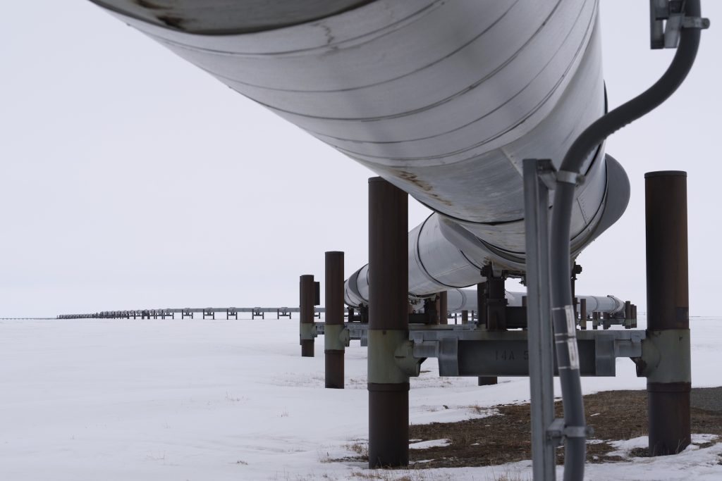 In the foreground, a close-up view of an above-ground pipeline. In the background, the pipeline extends to the horizon along a flat, snowy landscape.