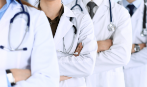 Four people in white lab coats with stethoscopes around their shoulders stand in a row with their arms crossed. Their faces are not visible.