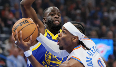 A basketball player in a white jersey attempts a shot while closely guarded by a defender in a blue and yellow Warriors uniform.