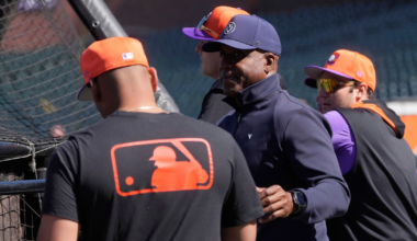Three men wearing baseball caps and casual sports gear stand near a batting cage, with one wearing a shirt featuring the MLB logo on the back.