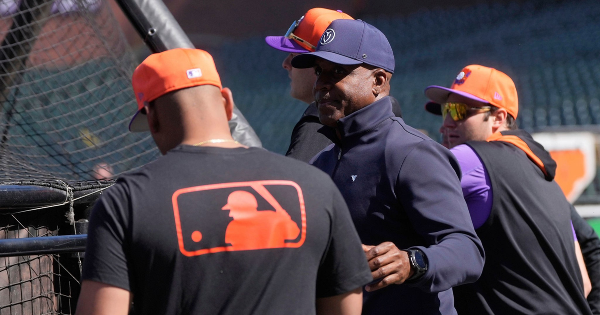 Three men wearing baseball caps and casual sports gear stand near a batting cage, with one wearing a shirt featuring the MLB logo on the back.