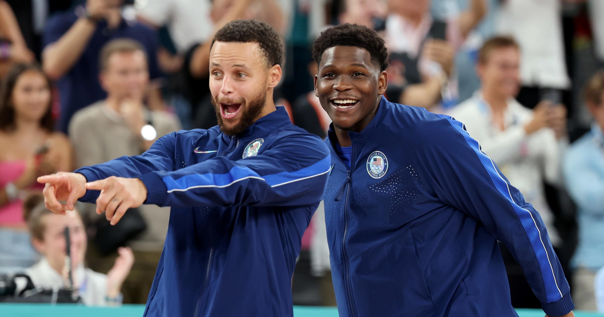 Two men in matching blue athletic jackets smile and gesture enthusiastically toward something off-camera, with a crowd blurred in the background.