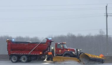 A red plow truck drives down a highway during a snowstorm.