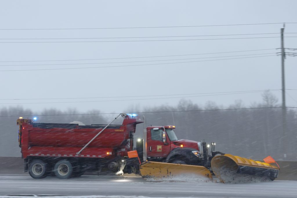 A red plow truck drives down a highway during a snowstorm.