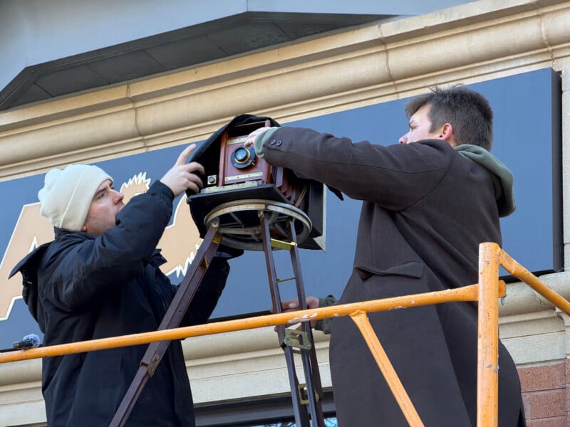 Two men stand on a scaffolding, adjusting an old-fashioned box camera mounted on a tripod outside a building. One is wearing a white beanie and dark jacket; the other is in a long brown coat.