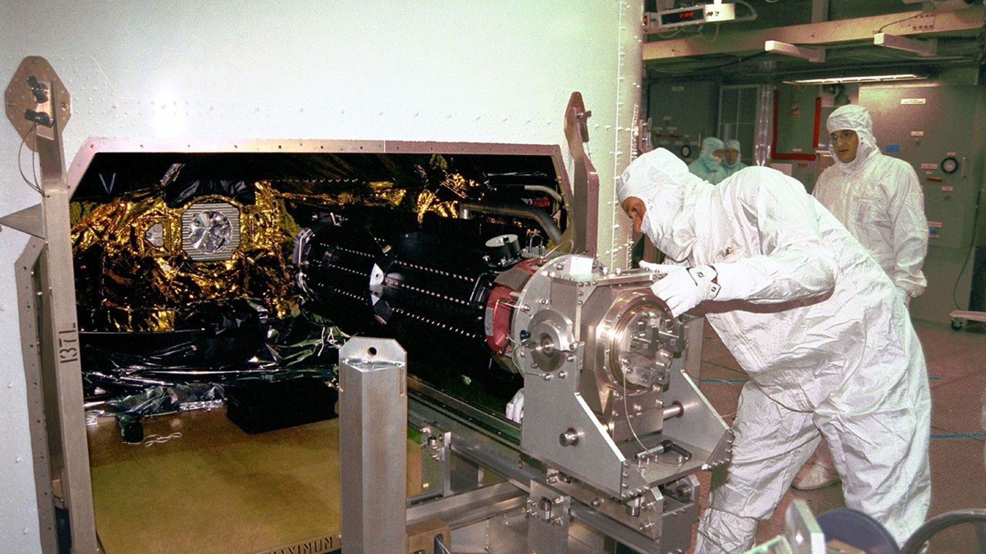 people in white clean suits load a metal cylinder into a large metal box covered in wires
