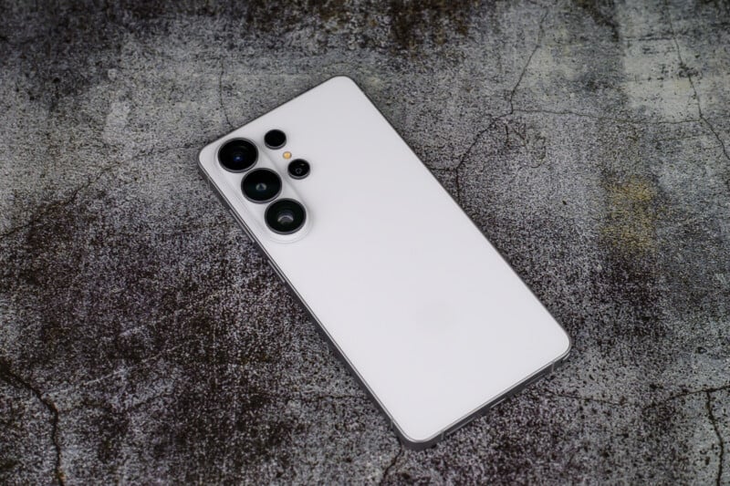 A white smartphone with three rear cameras and a flash lies face-down on a textured, gray concrete surface.