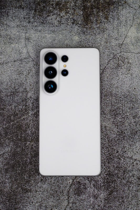 A white smartphone with four rear cameras and an LED flash is placed on a rough, dark textured surface. The Samsung logo is faintly visible on the back of the phone.