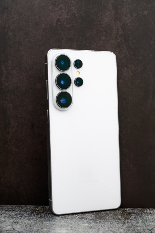 A white Samsung smartphone standing upright on a textured surface, showing its back with four camera lenses and an LED flash, set against a dark background.