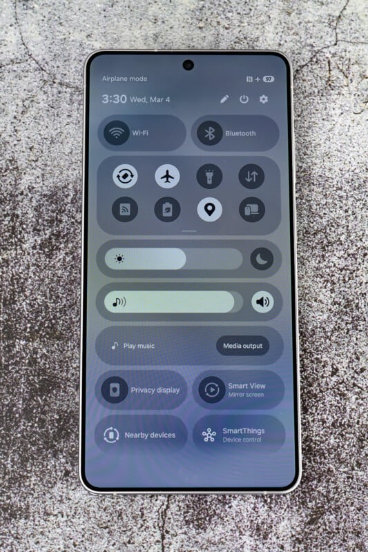 A smartphone displaying the quick settings menu, showing options like Wi-Fi, Bluetooth, Airplane mode, flashlight, location, and screen brightness slider. The phone is on a textured, concrete-like surface.