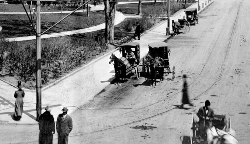 Black-and-white photo of a street scene with horse-drawn carriages, people walking, and some blurred motion. Utility poles, shadows, and trees are visible along the wide dirt road. The setting appears to be from the early 1900s.