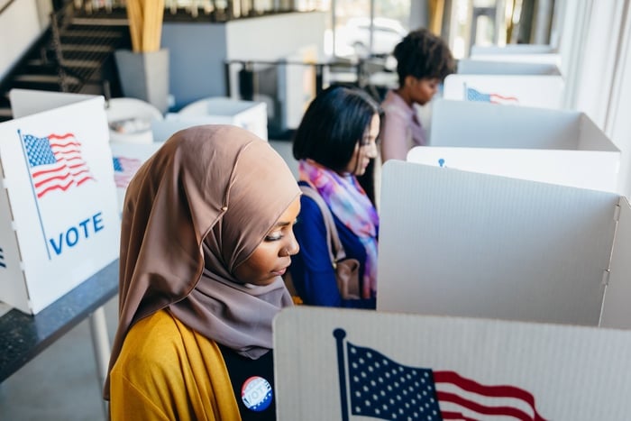 Three women stand at voting booths, each separated by partitions with American flags and the word "VOTE" on them. One woman in the foreground wears a hijab and a "VOTE" sticker. Sunlight shines through nearby windows.