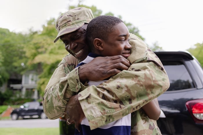 A smiling man in military uniform embraces a young boy outdoors, both appearing joyful and happy. A black vehicle and green trees are visible in the background.