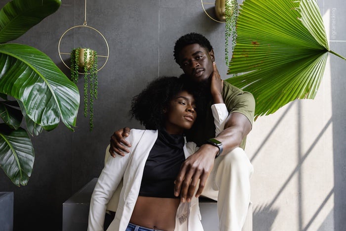 A stylish couple poses together indoors, surrounded by large green tropical plants and hanging planters. The woman leans back against the man, who has his arm around her shoulders. Natural light fills the space.