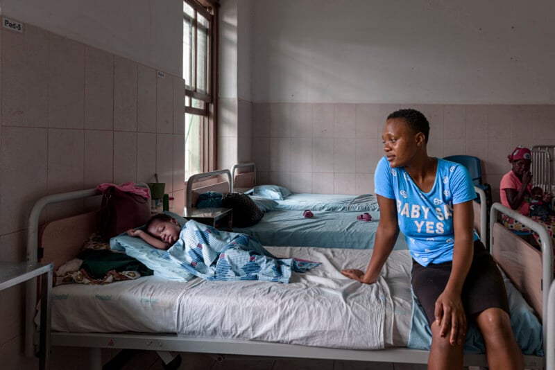 A woman sits on the edge of a hospital bed looking at a young girl who is lying under a blanket. In the background, another woman sits on a separate bed in a sparsely furnished hospital room.