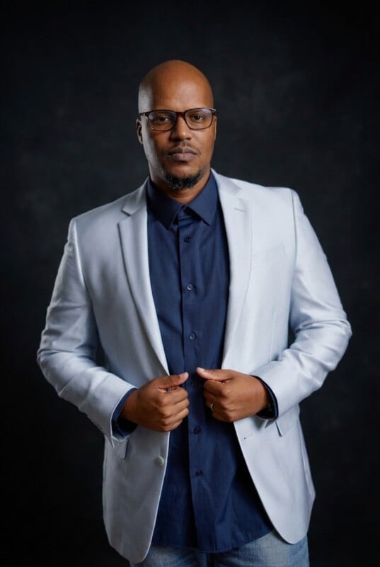 A man with glasses and a bald head wears a light gray blazer over a navy blue shirt. He stands against a dark background, facing forward with his hands adjusting his jacket.