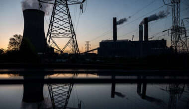 A bluish early dawn sky in the backdrop, with a dark set of buildings of a power plant, cooling towards, and transmission lines.