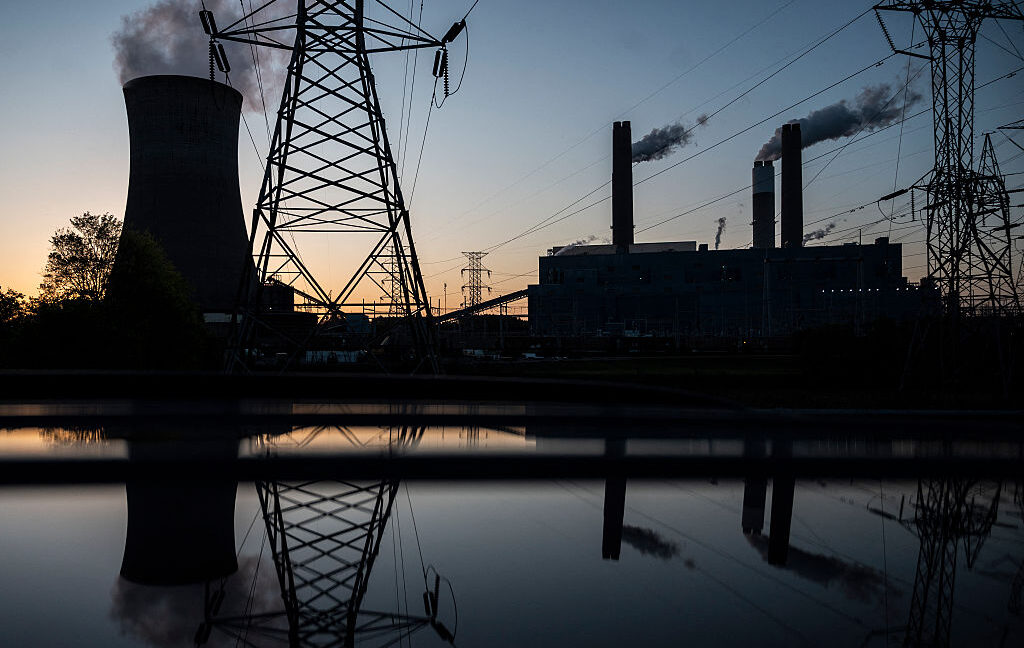 A bluish early dawn sky in the backdrop, with a dark set of buildings of a power plant, cooling towards, and transmission lines.