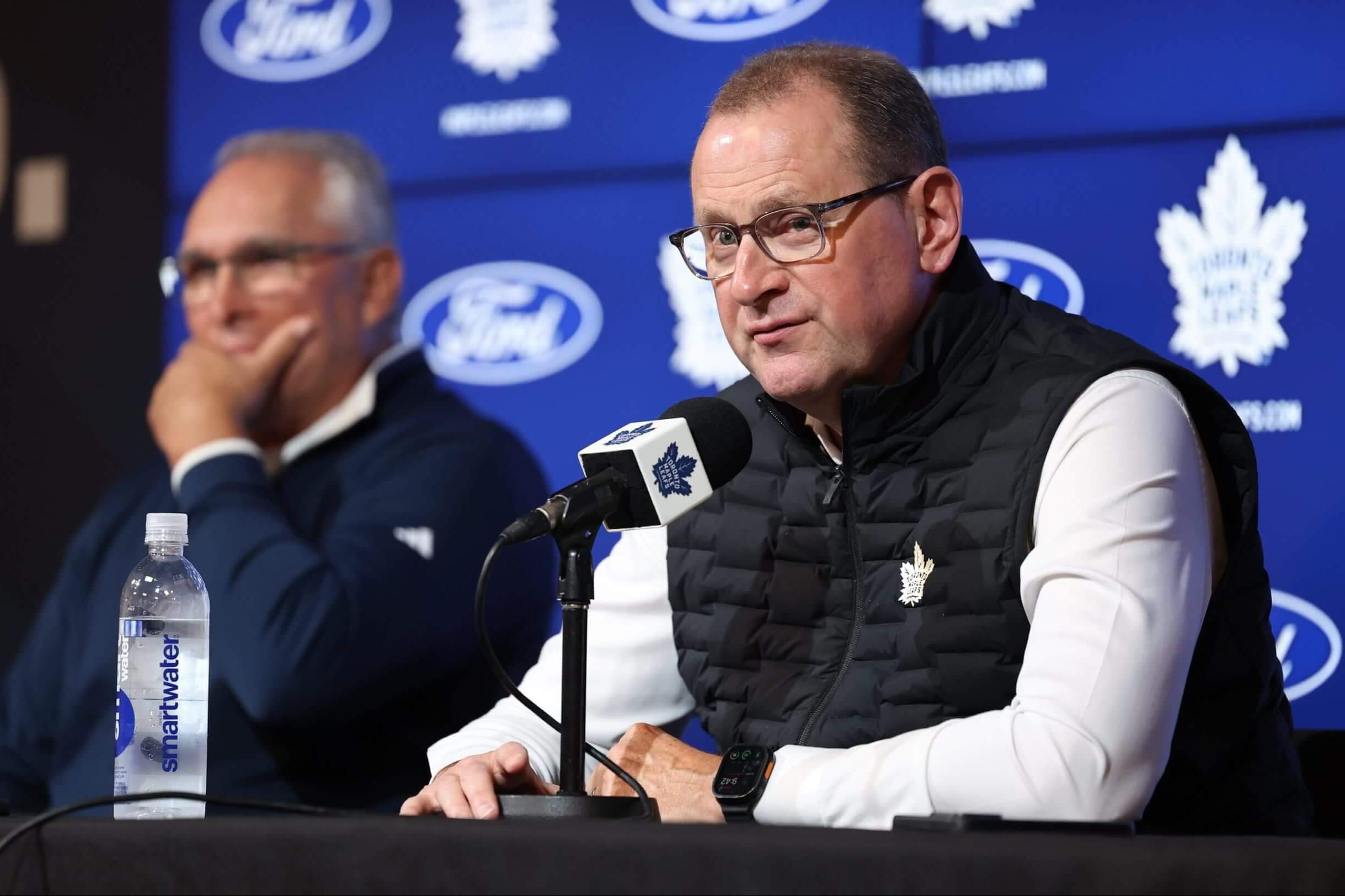 Brad Treliving answers questions at a press conference with Craig Berube in the background at the same table.
