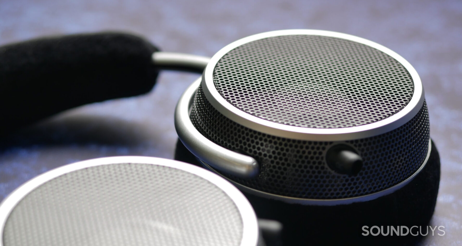 Close-up of Grell OAE2 open-back headphones earcup showing perforated metal grille, silver ring, and foam headband in soft focus background.
