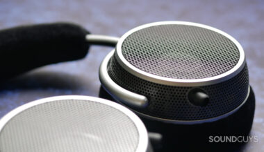Close-up of Grell OAE2 open-back headphones earcup showing perforated metal grille, silver ring, and foam headband in soft focus background.