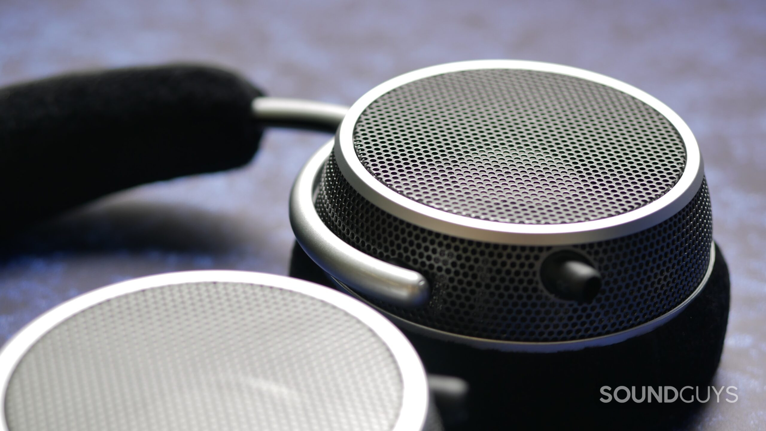 Close-up of Grell OAE2 open-back headphones earcup showing perforated metal grille, silver ring, and foam headband in soft focus background.