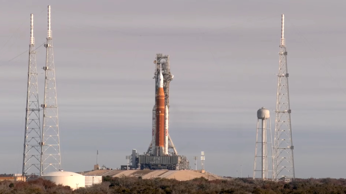 An orange rocket stands on a grey metal platform on a hill against a grey sky.