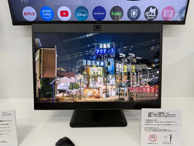A computer monitor displays a vibrant nighttime cityscape with illuminated Japanese signs and buildings. Streaming app icons like Netflix, Prime Video, and Disney+ are visible on a TV screen above the monitor.