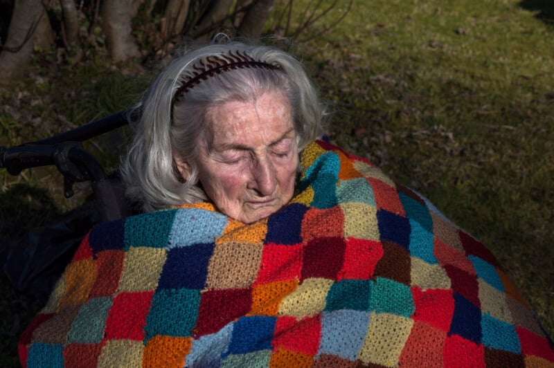 An elderly woman with long gray hair and a headband sits outdoors, wrapped in a colorful patchwork blanket, her eyes closed, with sunlight highlighting her face and the blanket.