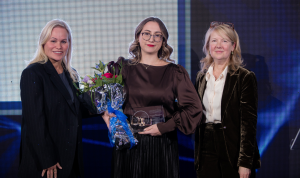 Three women stand on stage. The woman in the centre holds an award and a bouquet of flowers.