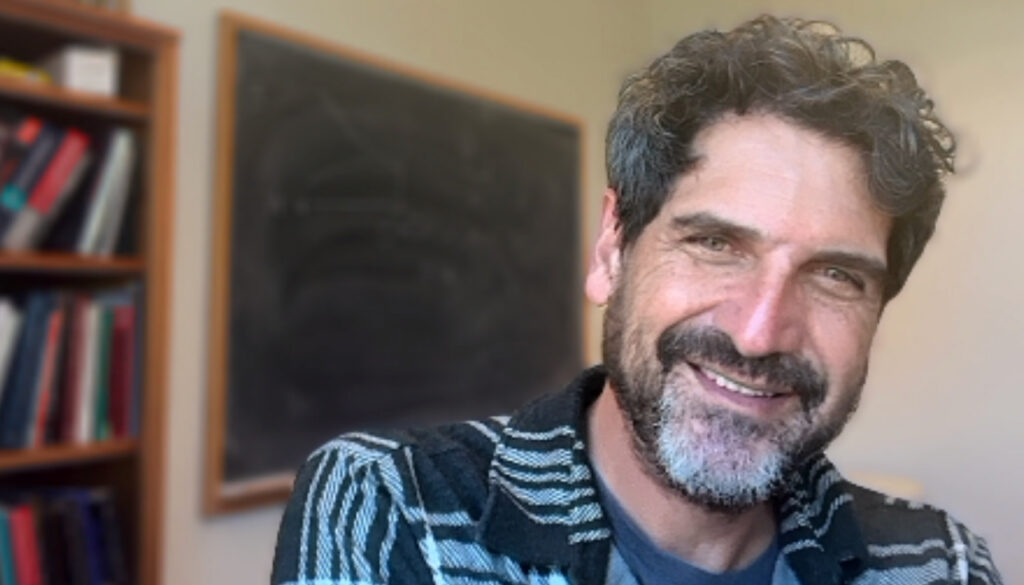 a bearded man smiling into the camera with a blackboard in the background