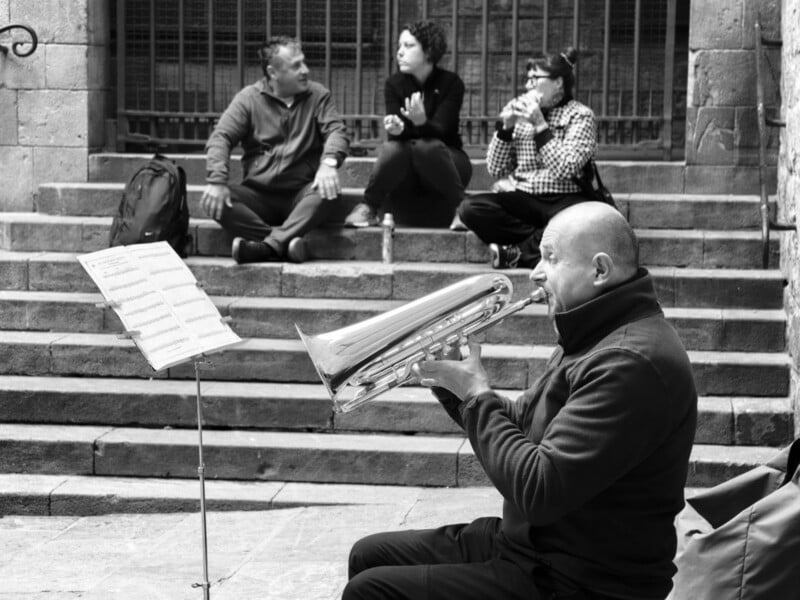A man plays a brass instrument while reading sheet music on a stand, seated on stone steps. Three people sit in the background, engaged in conversation, with a metal fence and stone wall behind them.