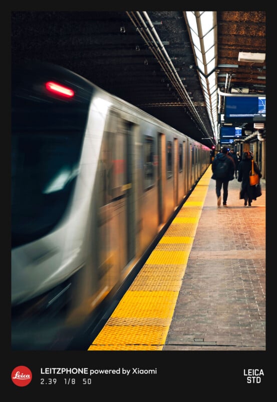 A subway train speeds into an underground station as blurred commuters walk along the yellow safety platform. Overhead lights illuminate the scene, and signs hang from the ceiling. The image has camera settings and branding at the bottom.