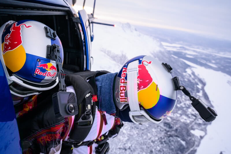Two skydivers wearing Red Bull helmets and jumpsuits prepare to jump from a helicopter over a snowy, mountainous landscape, both equipped with cameras on their helmets.