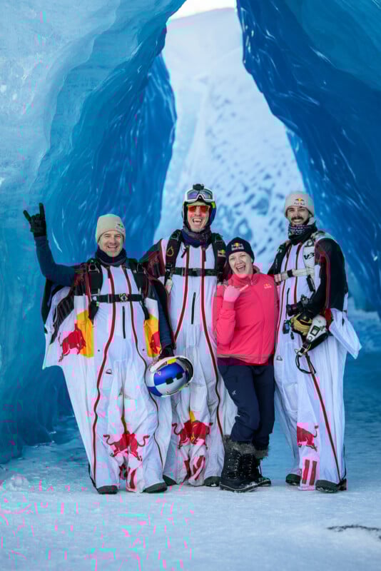 Four people in cold-weather gear pose in a blue ice cave. Three wear white wingsuits with red bull logos, holding helmets, while the fourth wears a pink top and black pants, smiling and gesturing with one hand.