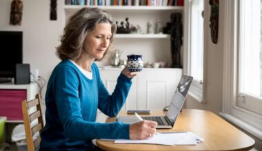 Mature Caucasian woman sat at a table with coffee and laptop while making notes on paper