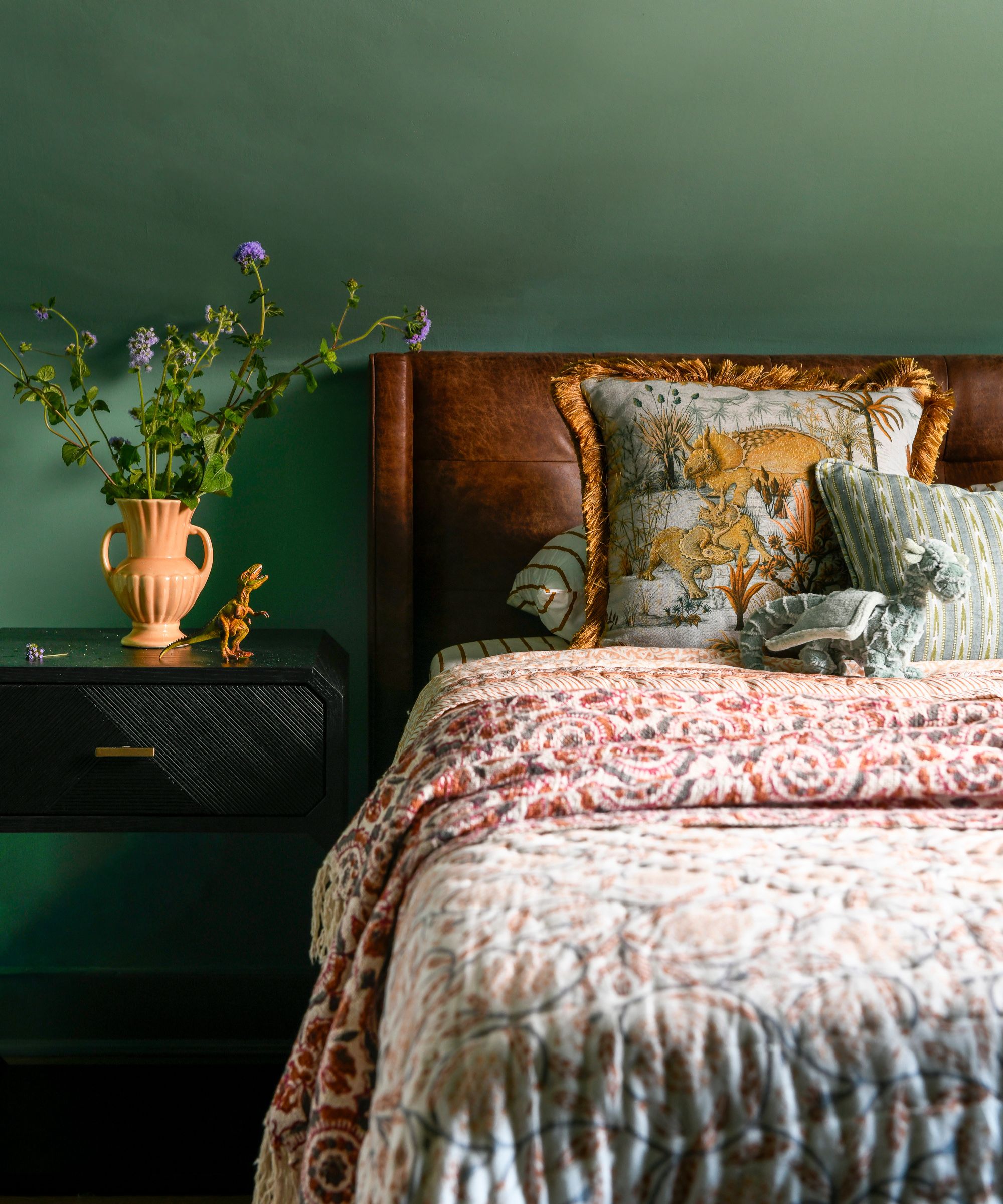 A bedroom with dark green walls, patterned bedding and cushions, and a black nightstand with a vase of flowers.