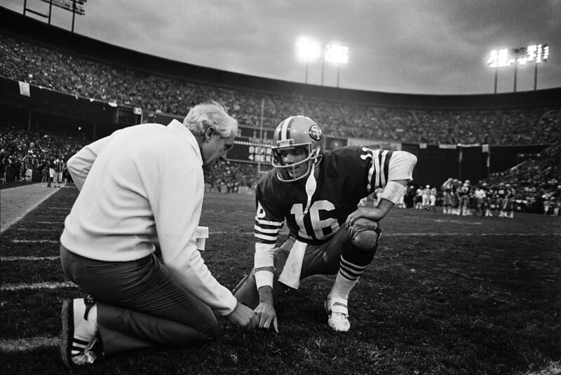 A football coach kneels on the sideline talking to a player in a number 16 San Francisco 49ers uniform during a game, with a crowded stadium in the background.