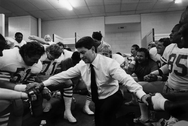 A football coach in a suit kneels in a locker room, holding hands with players in uniform as they bow their heads in a pre-game huddle, creating a moment of unity and focus.