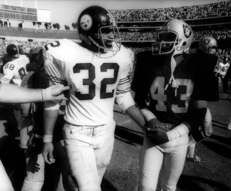 Two American football players from opposing teams, one in a white jersey (number 32) and one in a dark jersey (number 43), shake hands on the field after a game, with a stadium full of spectators in the background.
