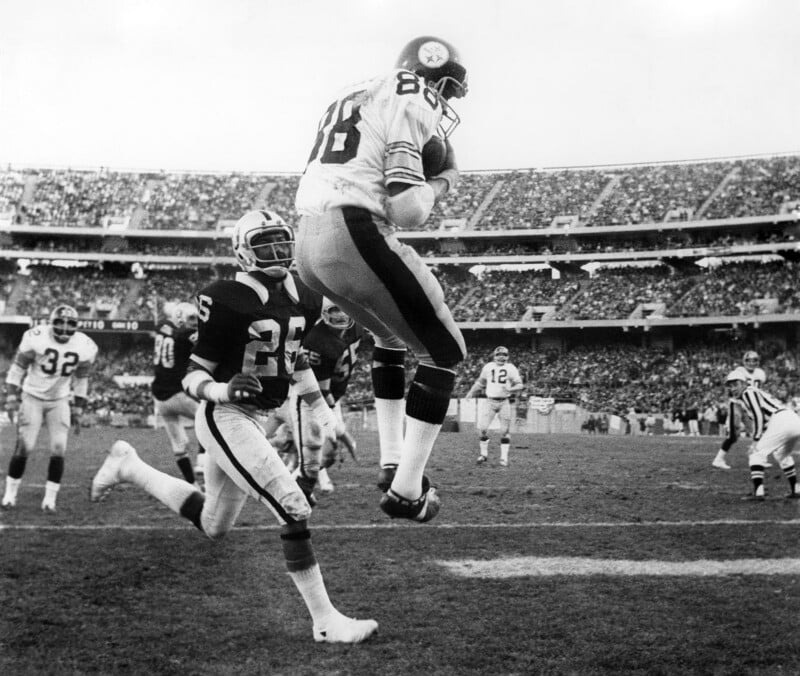 A football player in a white jersey leaps to catch a pass in the end zone as a defender in a dark jersey pursues him during an outdoor game, with a crowded stadium in the background.