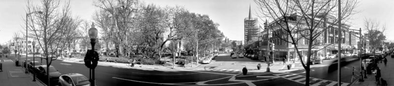 A wide, black-and-white panoramic view of a city intersection with trees, pedestrians, cars, and several multi-story buildings lining the streets. The sky is clear, and leafless branches frame the scene.