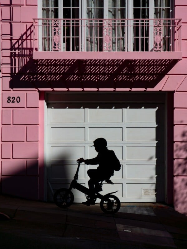A person wearing a helmet and backpack rides a bicycle past a white garage door on a pink building. Shadows and sunlight create a silhouette effect. The number 820 is visible on the wall.