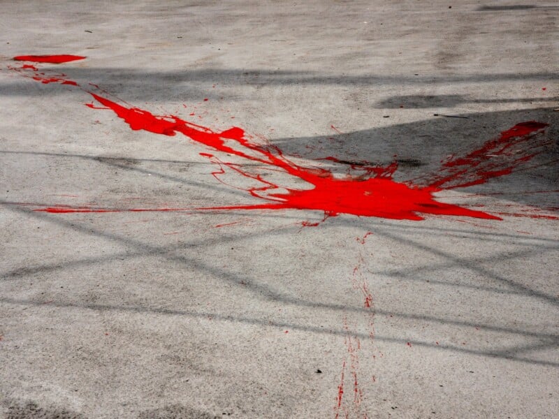 Bright red paint is splattered across a rough, gray concrete surface, creating irregular shapes and streaks. Shadows of a metal fence or structure are cast over the area.