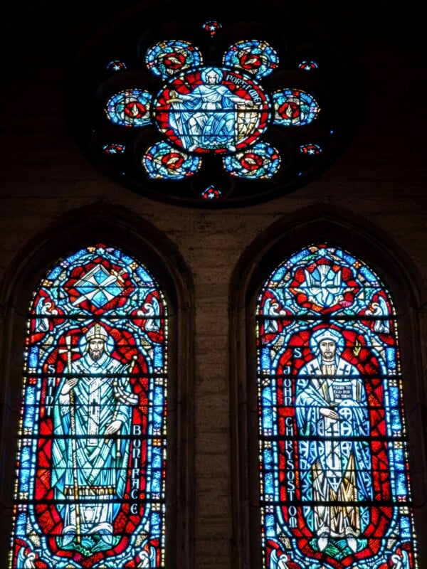 Two tall stained glass windows with religious figures beneath a circular stained glass panel featuring a central seated figure and intricate patterns, set in a stone wall. The glass is detailed with blue, red, and white colors.