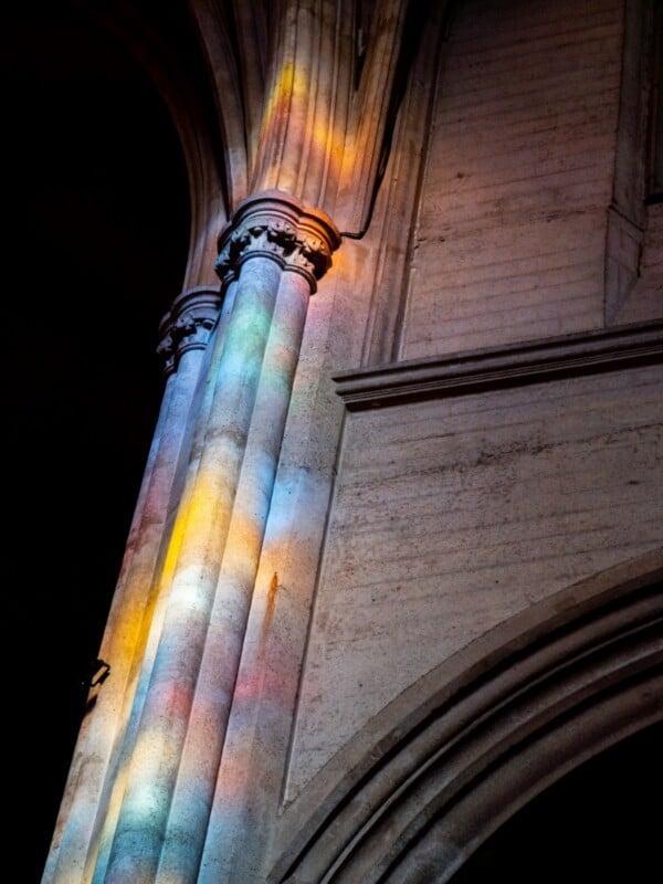 Colored light from stained glass windows casts rainbow hues onto a tall stone column inside a cathedral, highlighting the architectural details and texture of the pillar.